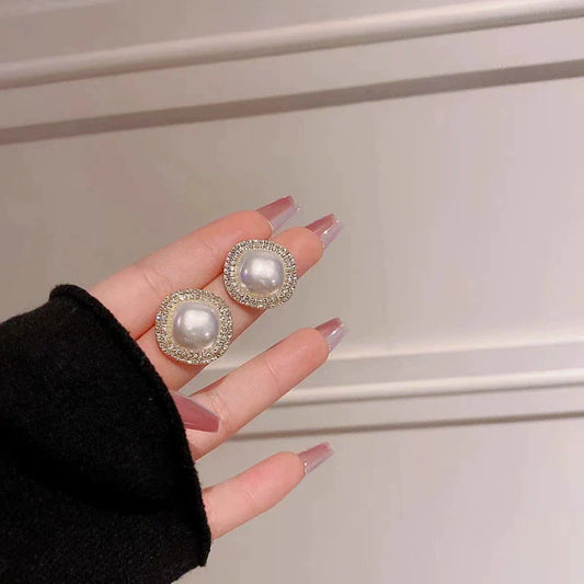 Pearl-like earrings held by a hand with black sleeve and pink nails against a neutral background